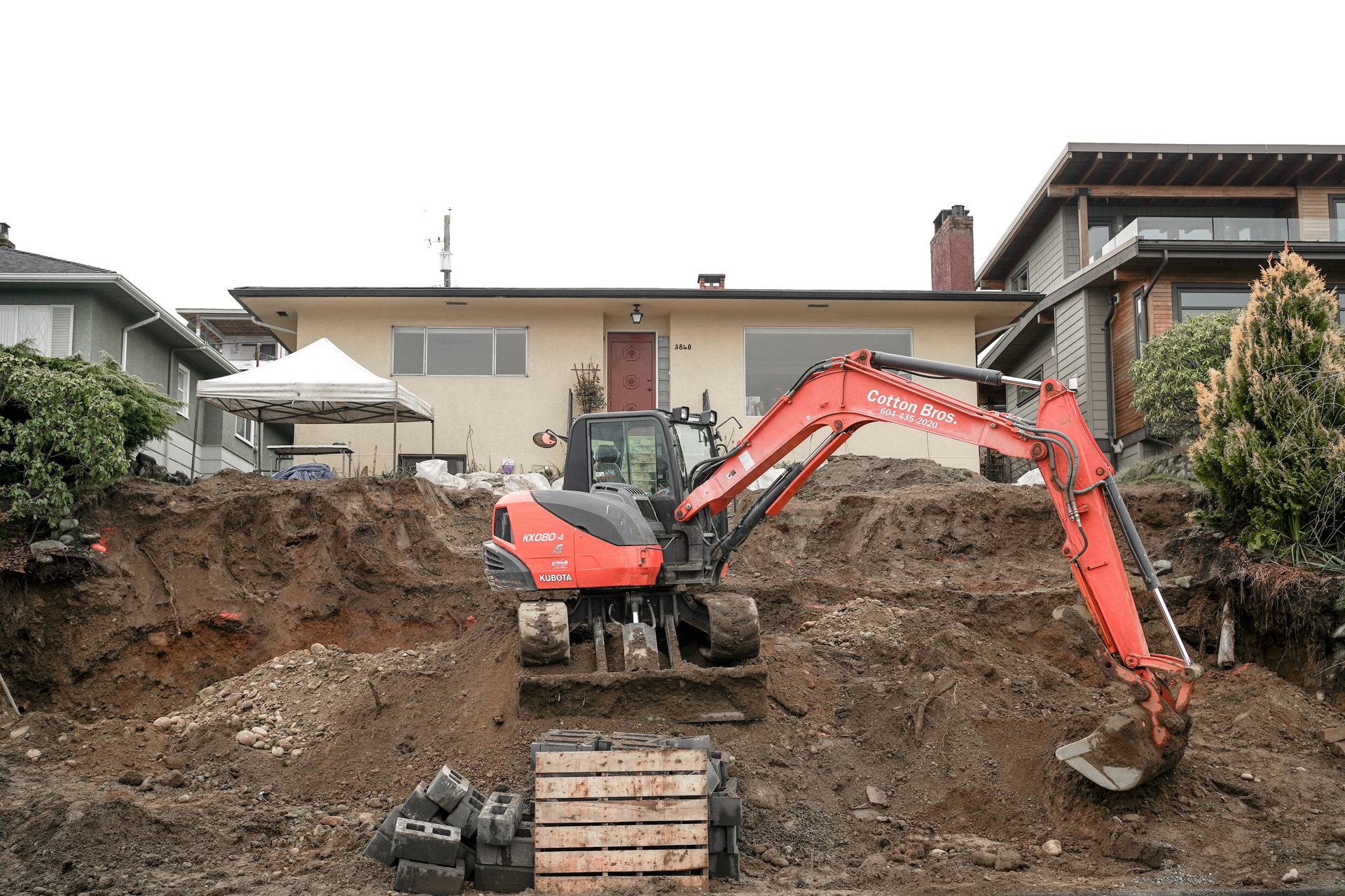 a red excavator digging a pile of dirt in front of a house